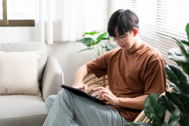Teknoloji kavramı. Günlük tişörtü ve kotuyla rahat bir şekilde tahta koltukta oturan ve iPad 'deki web tarayıcısını kontrol etmek için dokunmatik ekran yapan erkek..