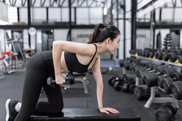 exercise concept The female exercise beginner doing dumbbell workout by resting her left knee and left hand on the bench and raise her right elbow up with a dumbbell in her hand.
