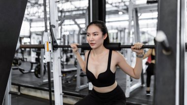 Egzersiz konsepti siyah spor elbiseli kadın ağırlık makinesini omuzlarında taşıyıp kaldırmaya çalışıyor..