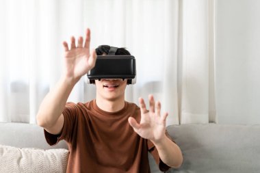 Teknoloji kavramı. Kahverengi tişörtü ve açık mavi kotlu adam koltukta otururken sanal gerçeklik kulaklığı takıyor..