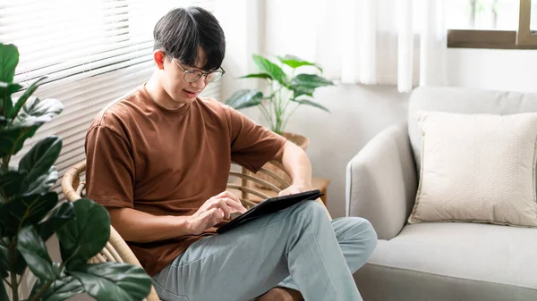 Teknoloji kavramı. Günlük tişörtü ve kotuyla rahat bir şekilde tahta koltukta oturan ve iPad 'deki web tarayıcısını kontrol etmek için dokunmatik ekran yapan erkek.