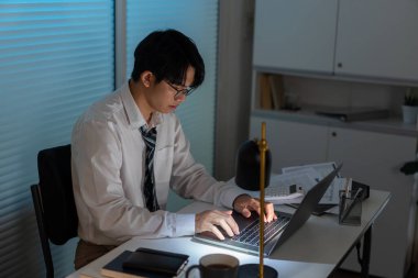 Fazla mesai. Ofis personeli masasında oturmuş, evrak okuyor ve not defterine yazı yazıyor.