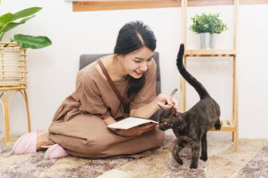Evcil hayvan sevici kavramı, kediyle oynayan Asyalı genç bir kadın ve oturma odasındaki not defteri..