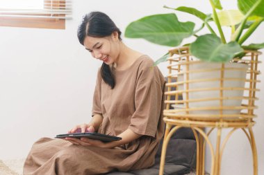 Rahatlama yaşam tarzı konsepti, oturma odasında sosyal medyada sörf yapmak için tablet kullanan genç Asyalı kadın..