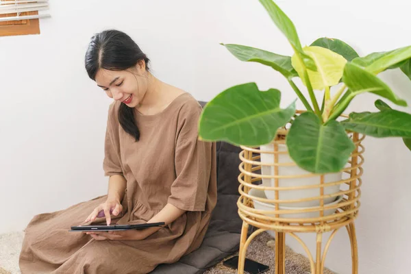 Rahatlama yaşam tarzı konsepti, oturma odasında sosyal medyada sörf yapmak için tablet kullanan genç Asyalı kadın..
