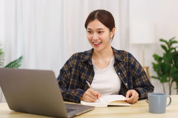 客厅概念中的生活方式 年轻的亚洲女人使用笔记本电脑 并在笔记本上记录数据 — 图库照片