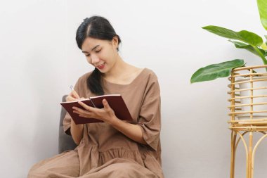 Rahatlama yaşam tarzı konsepti, genç Asyalı kadının oturma odasında otururken deftere yazı yazması..