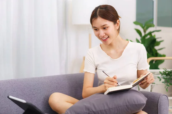 客厅里的生活方式 年轻的亚洲女人在看平板电脑 在笔记本上写数据 — 图库照片