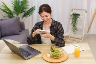 Oturma odası konseptinde yaşam tarzı, akıllı telefon kullanan ve sebze salatası yiyen genç Asyalı kadın..