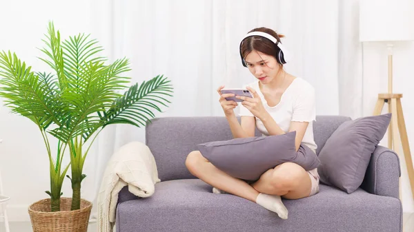 Lifestyle in living room concept, Young Asian woman in headphone to playing games on smartphone.