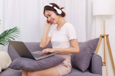 Oturma odasındaki yaşam tarzı, dizüstü bilgisayardan müzik dinleyen kulaklıklı genç Asyalı kadın..