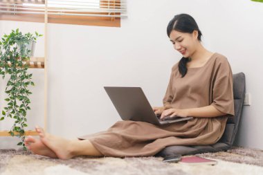 Rahatlama yaşam tarzı konsepti, oturma odasında sosyal medyada sörf yapmak için dizüstü bilgisayar kullanan genç Asyalı kadın..