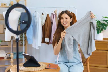 Online shopping concept, Young Asian women hold clothes to present and selling on video streaming.
