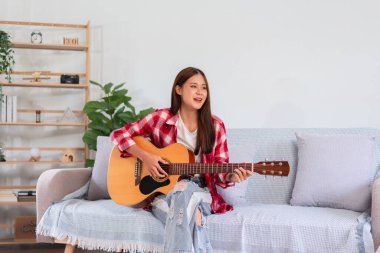 Müzikle rahatlama kavramı, genç Asyalı kadın kanepede şarkı söylemek için akustik gitar çalıyor..
