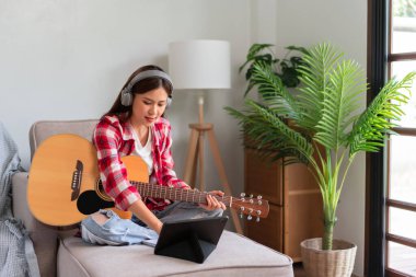 Gitar ve şarkıcı konsepti. Tablette müzik öğrenirken akustik gitar çalan Asyalı genç bir kadın..