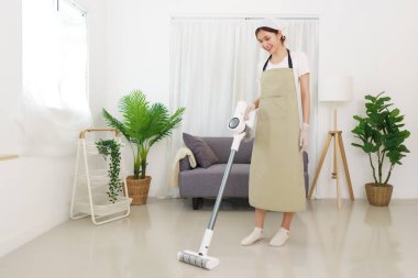 Oturma odası konseptinde yaşam tarzı, yerleri temizlemek için elektrikli süpürge kullanan genç Asyalı kadın..