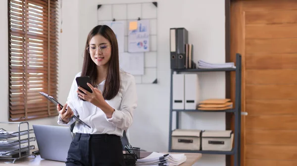 Business concept, Businesswoman holding clipboard and stand to reading business data in smartphone.