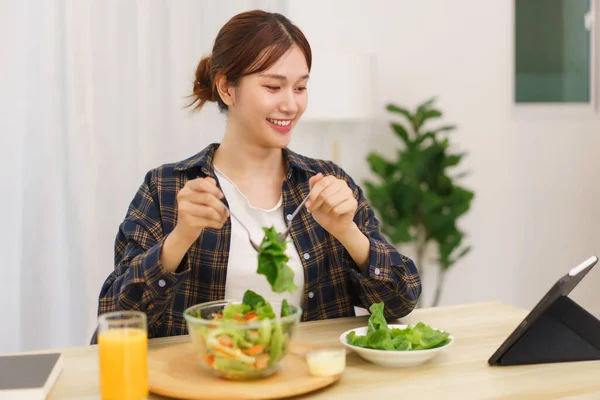 Lifestyle in living room concept, Young Asian woman looking at tablet and eating vegetable salad.