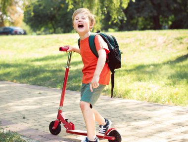 Küçük çocuk ağlıyor. O bir scooter tutuyor. Kırmızı tişörtlü yeşil çimenli bir çocuk.