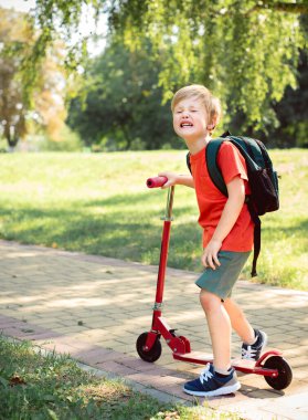 Küçük çocuk ağlıyor. O bir scooter tutuyor. Kırmızı tişörtlü yeşil çimenli bir çocuk.