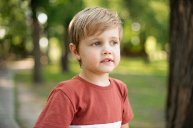 Küçük ciddi çocuk. Yeşil Park 'ın arka planında duruyor. Çocuk başka tarafa bakıyor.