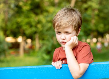 Mavi çitin yanındaki yeşil parkın arka planında oturan yakışıklı bir çocuğun portresi. Adam düşünceli bir şekilde yana bakıyor.