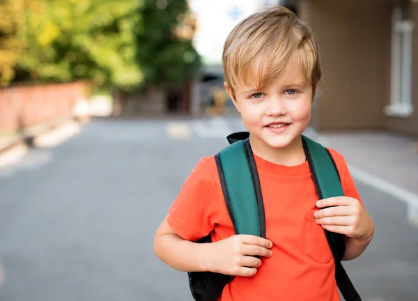 Niño sonriente con una camiseta roja y una mochila. Está parado en la calle. — Foto de Stock