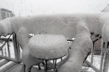Bulutlu bir günde pencere camına buz damlaları, seçici bir odak. Teras manzarası, karla kaplı masa ve kışın pencereden sandalyeler. Doğal afetler, dondurucu yağmur, don, yoğun kar yağışı. 