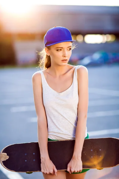 Primer plano retrato de verano de una mujer joven y bonita posando en la ciudad, chica hipster con tabla de skate en la ciudad. Verano noche luz del sol — Foto de Stock