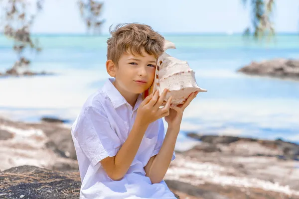 Un garçon tenant et entendant un énorme gros coquillage près de l'oreille sur la plage tropicale d'été — Photo