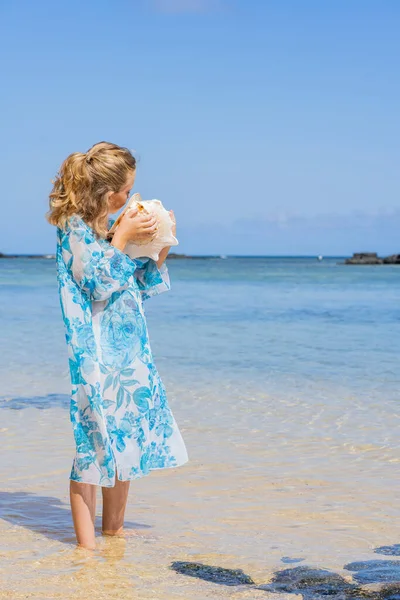 Ένα κορίτσι που κρατά και ακούει τεράστια κοχύλια κοντά στο αυτί στην καλοκαιρινή τροπική παραλία — Φωτογραφία Αρχείου