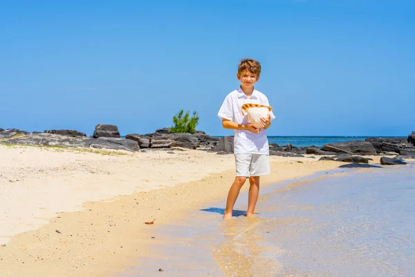 Un ragazzo che cammina con una grande conchiglia in mano sulla spiaggia tropicale. — Foto Stock