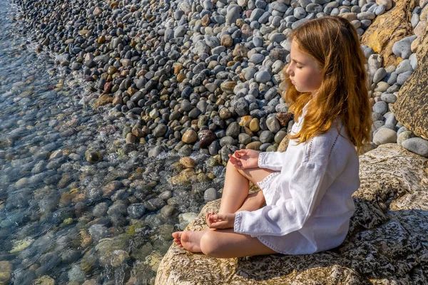 Deniz kenarındaki bir kayanın üzerinde oturmuş meditasyon yapan güzel küçük bir kız. Yüksek kalite fotoğraf