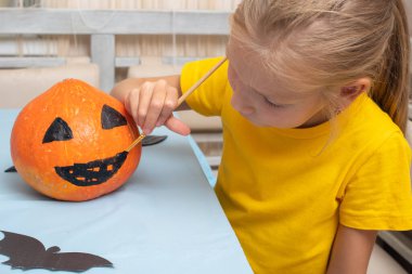 Tatlı genç bir kız oturma odasındaki masada oturuyor, turuncu bir balkabağına korkunç bir yüz çiziyor, fırçayla.