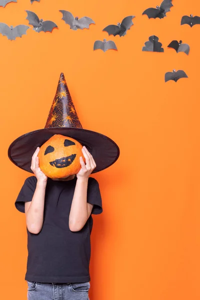 Niño en un sombrero sosteniendo una calabaza de halloween delante de su cabeza sobre un fondo naranja con murciélagos de papel — Foto de Stock