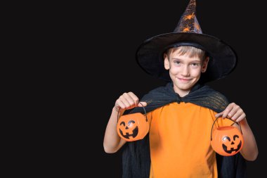 Büyücü şapkalı ve turuncu tişörtlü çocuk Cadılar Bayramı balkabağı şeklinde kovalarca şeker tutuyor.