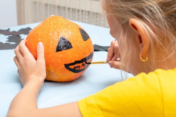 Χαριτωμένο κορίτσι ζωγραφική τρομακτικό πρόσωπο σε πορτοκαλί κολοκύθα με πινέλο για διακόσμηση στο σπίτι κατά τη διάρκεια των διακοπών Halloween — Φωτογραφία Αρχείου