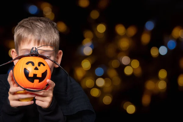 Ett barn som håller en orange pumpa-formad korg med ett leende ansikte och ett slagträ på en svart bakgrund med bokeh — Stockfoto