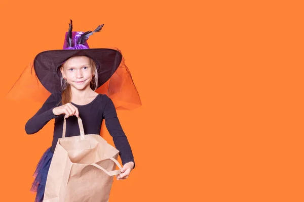 Une belle fille souriante dans une robe noire et un chapeau de sorcière ouvre un sac en papier artisanal sur un fond orange. — Photo