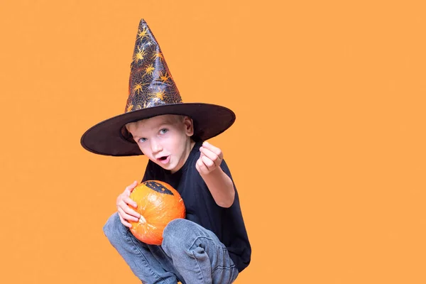 Les enfants Halloween. Petit garçon émotionnel dans un chapeau de sorcier tenant une citrouille orange dans ses mains et montrant un bonbon — Photo