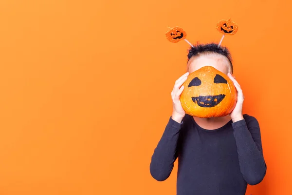 Halloween dzieci. Dziewczyna trzymająca dynię przed twarzą z pomalowaną przerażającą twarzą na pomarańczowym tle — Zdjęcie stockowe