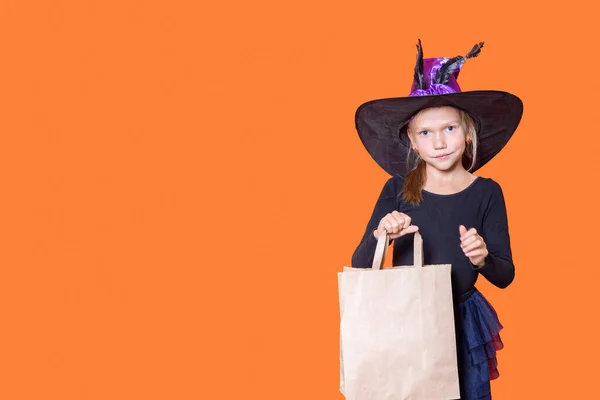 Une belle fille en robe noire et un chapeau de sorcière tient un sac en papier artisanal, regardant la caméra et souriant, sur une orange — Photo