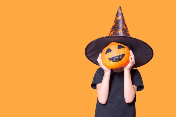 Chłopiec w kapeluszu czarodzieja trzymający dynię przed twarzą z pomalowaną przerażającą twarzą na pomarańczowym tle. — Zdjęcie stockowe