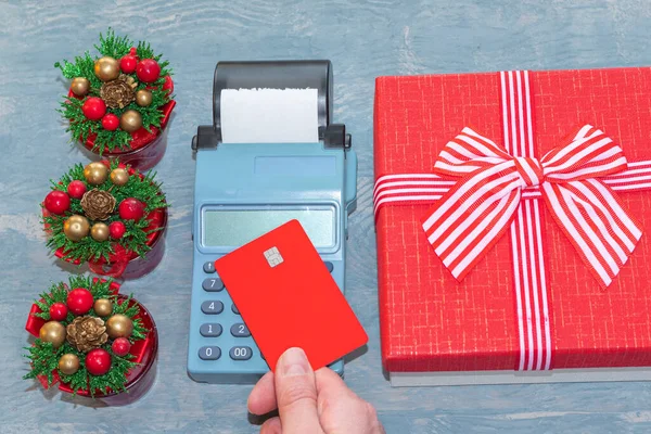 Рука с красной банковской карточкой над кассой рядом с красной подарочной коробкой с ленточкой и маленькими елками — стоковое фото
