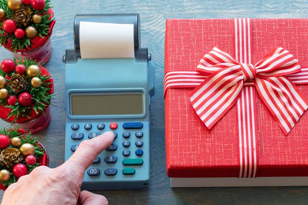 Una mano presionando los botones de la caja registradora junto a una caja de regalo roja con una cinta y pequeños árboles de Navidad — Foto de Stock