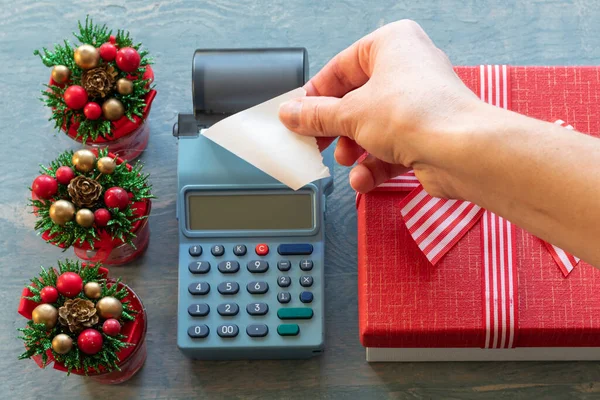 Venta de Navidad. Mano sosteniendo Un cheque blanco en blanco de una caja registradora cerca de una caja de regalo roja con una cinta y pequeños árboles de Navidad con conos. Comprar regalos para Año Nuevo y Navidad — Foto de Stock