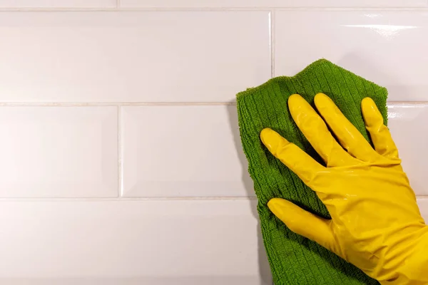 Hand in Hand wischt ein gelber Gummihandschuh mit einem grünen Lappen die Keramikfliesenoberfläche ab, Nahaufnahme, Kopierraum. Desinfektion der Küche, Bad. Hausputzkonzept — Stockfoto