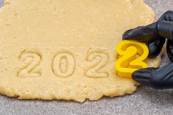 Uma mão em uma luva preta cortando a data 2022 da massa crua de Ano Novo e biscoitos de Natal — Fotografia de Stock