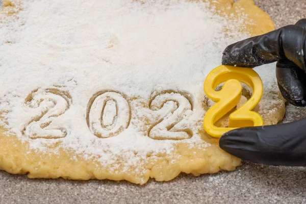 Uma mão em uma luva preta que corta a data 2022 de biscoitos de Ano Novo da massa de farinha crua salpicada de farinha branca — Fotografia de Stock