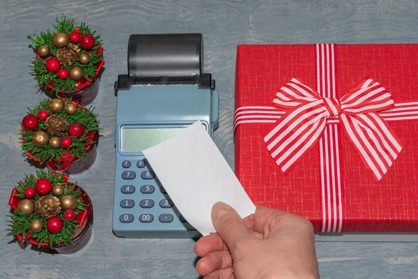 Venta de Navidad. Una mano sosteniendo un cheque blanco en blanco encima de la caja registradora cerca de una caja de regalo roja con una cinta y pequeños árboles de Navidad con conos. Comprar regalos para Año Nuevo y Navidad — Foto de Stock